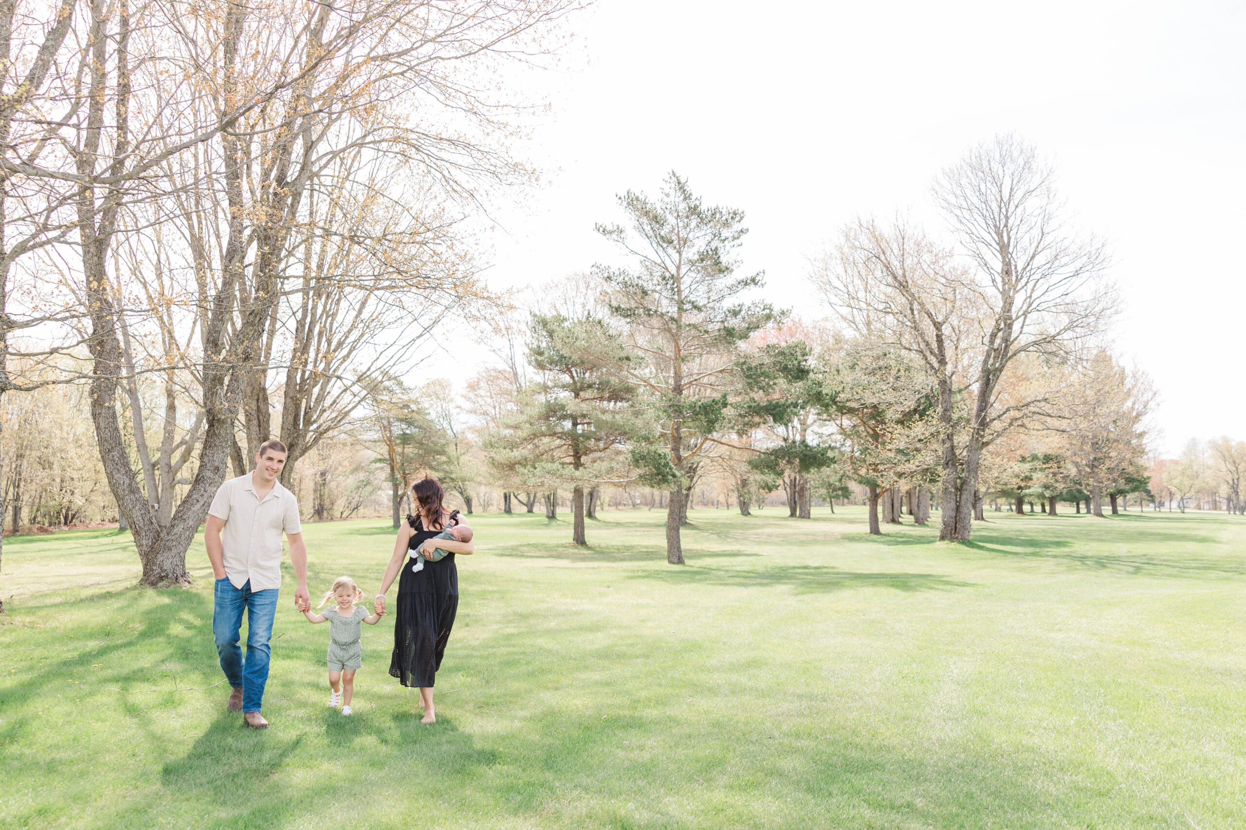 Family walking hand in hand through a sunlit park during their spring newborn photography session in Northern Michigan