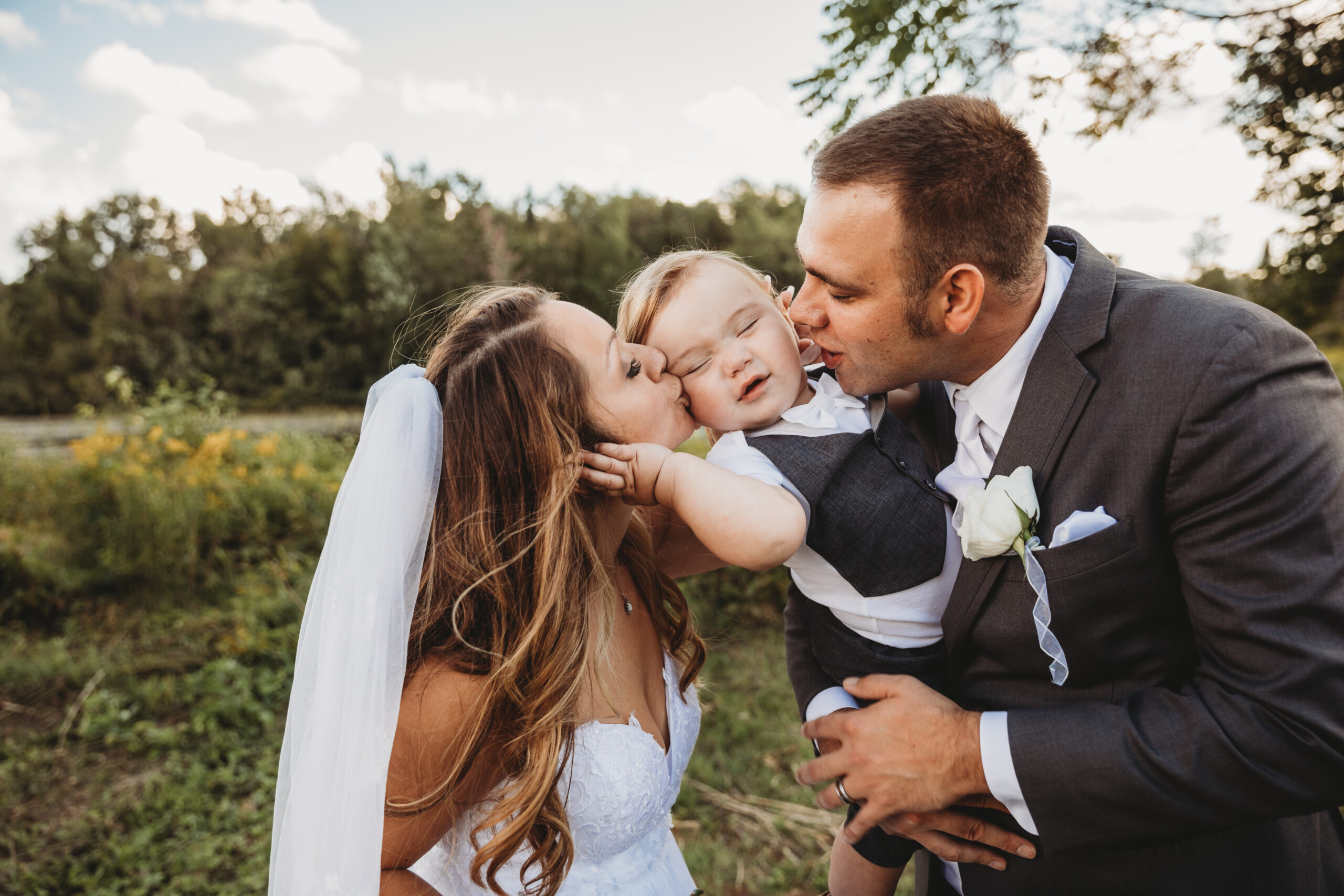 Bride and groom kissing their young son on the cheek — a joyful family moment on their wedding day