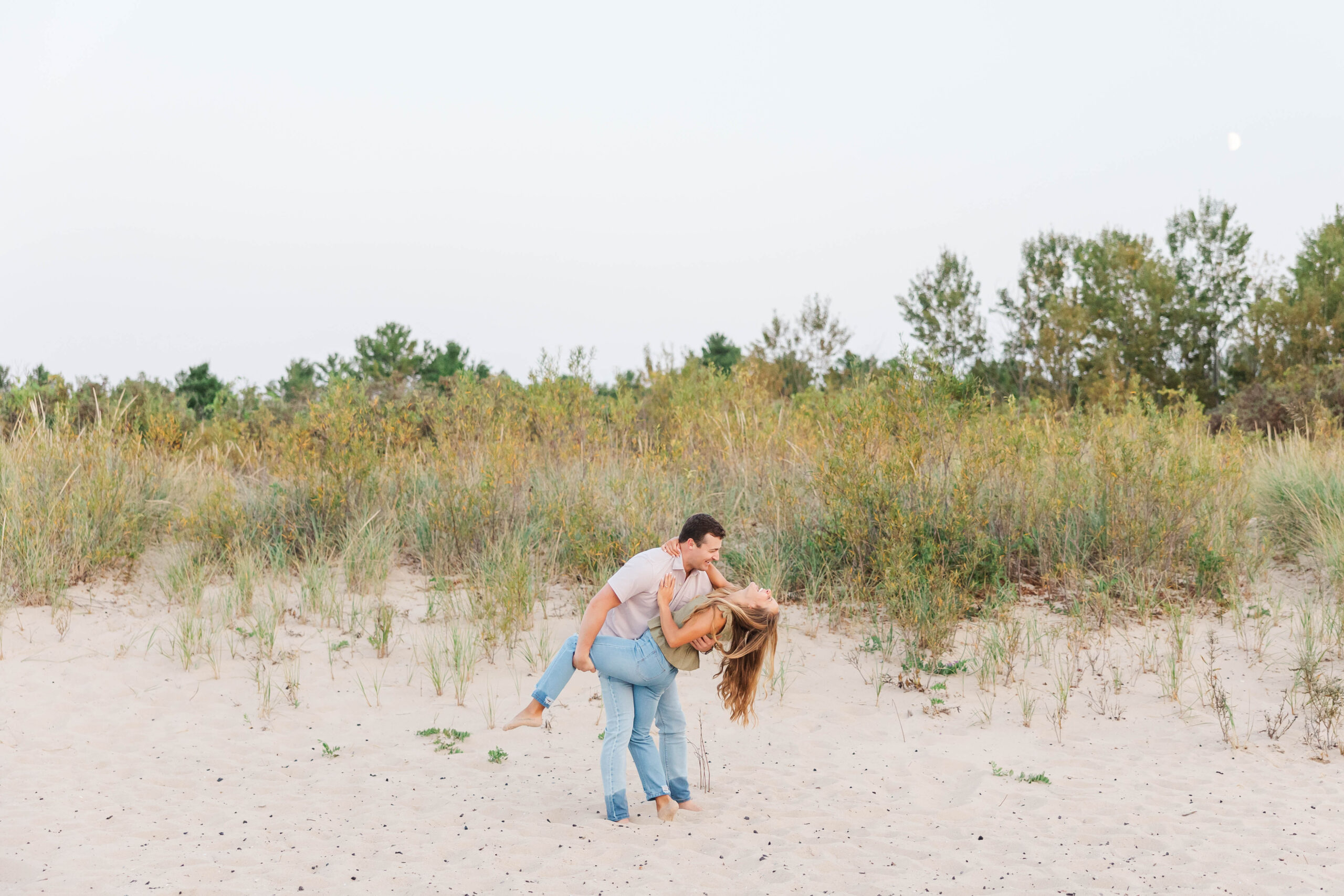 Playful couple during their beach engagement session in Northern Michigan wearing casual jeans and neutral tops