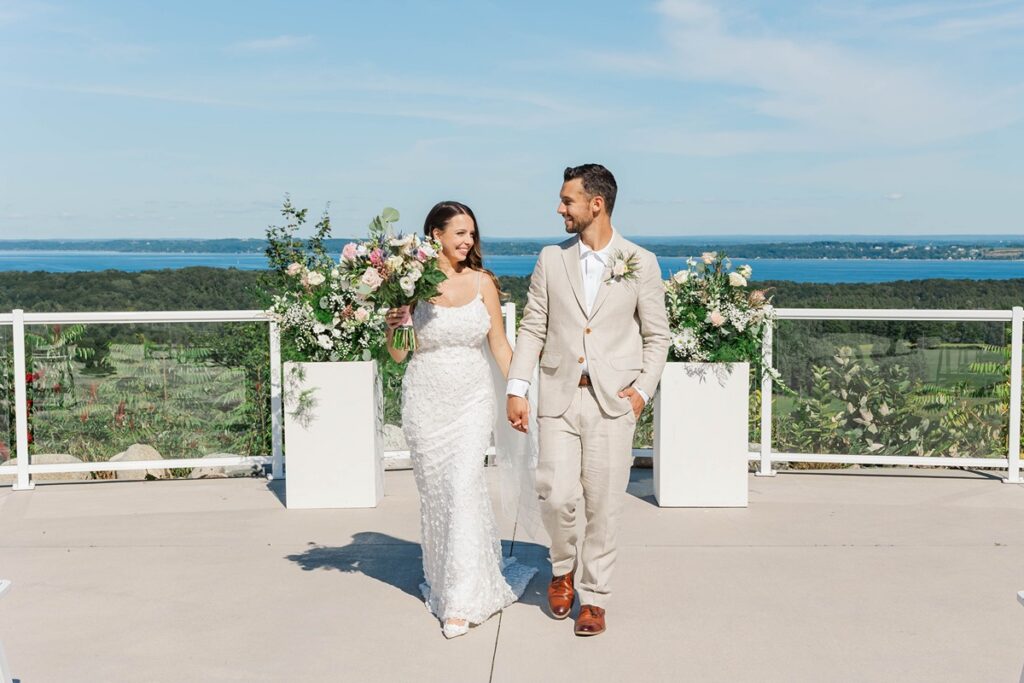 Bride and groom walking hand in hand at Bayview Weddings at Gallagher Farms in Cedar, Michigan, overlooking Grand Traverse Bay during a luxury vineyard wedding.