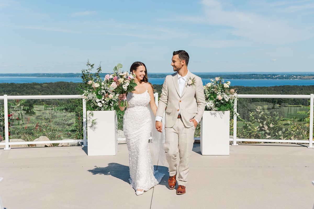 Bride and groom walking hand in hand at Bayview Weddings at Gallagher Farms in Cedar, Michigan, overlooking Grand Traverse Bay during a luxury vineyard wedding.
