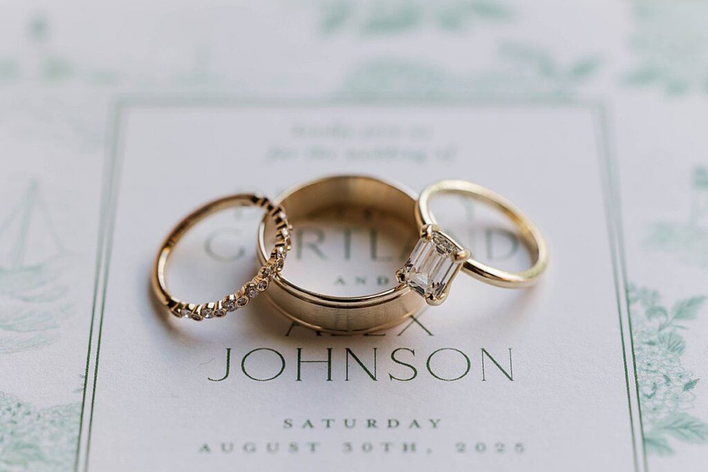 gold engagement ring and wedding bands stacked on invitation suite for wedding detail photos at Bayview Weddings at Gallagher Farms