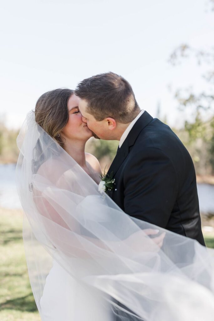 Madison and Jaxon sharing a kiss beneath her flowing veil during portraits at their Mill Pond Event Center wedding in Kalkaska, Michigan.