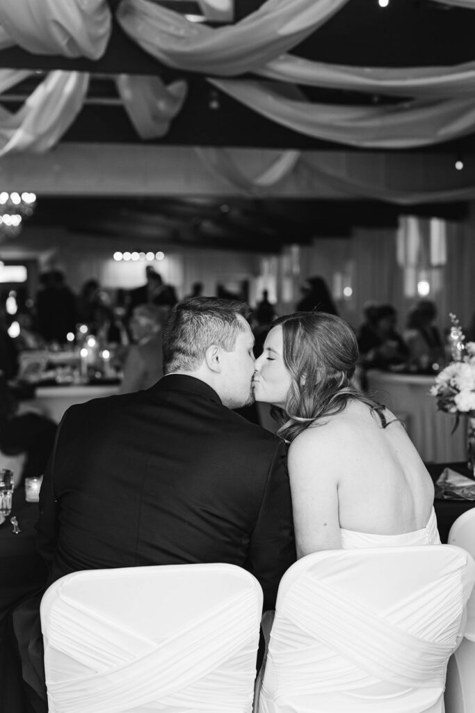 Candid black-and-white photo of Madison and Jaxon sharing a kiss during their wedding reception at Mill Pond Event Center in Kalkaska, Michigan.