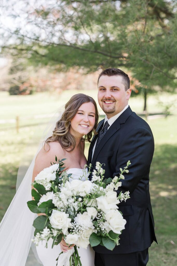 Bride and groom smiling together with the bride's white bouquet during their Mill Pond Event Center wedding in Kalkaska, Michigan.