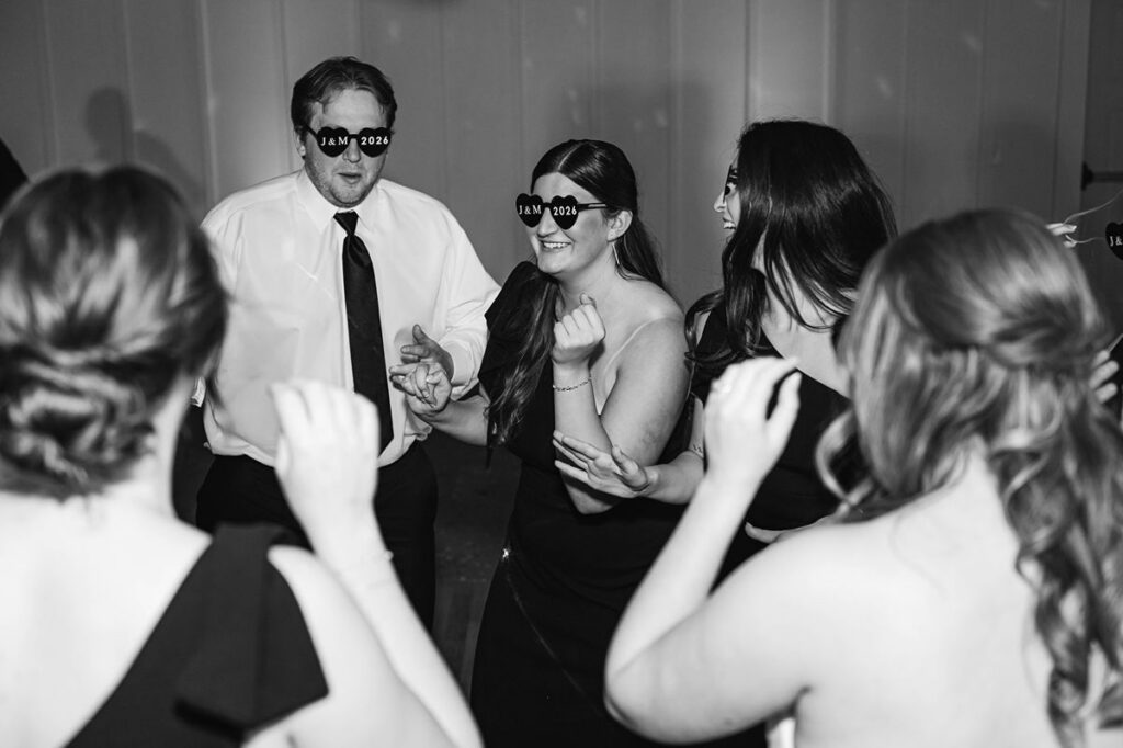 Documentary-style black-and-white dance floor photo of wedding guests celebrating in custom J&M 2026 party glasses during Madison and Jaxon's Mill Pond Event Center wedding reception in Kalkaska, Michigan.