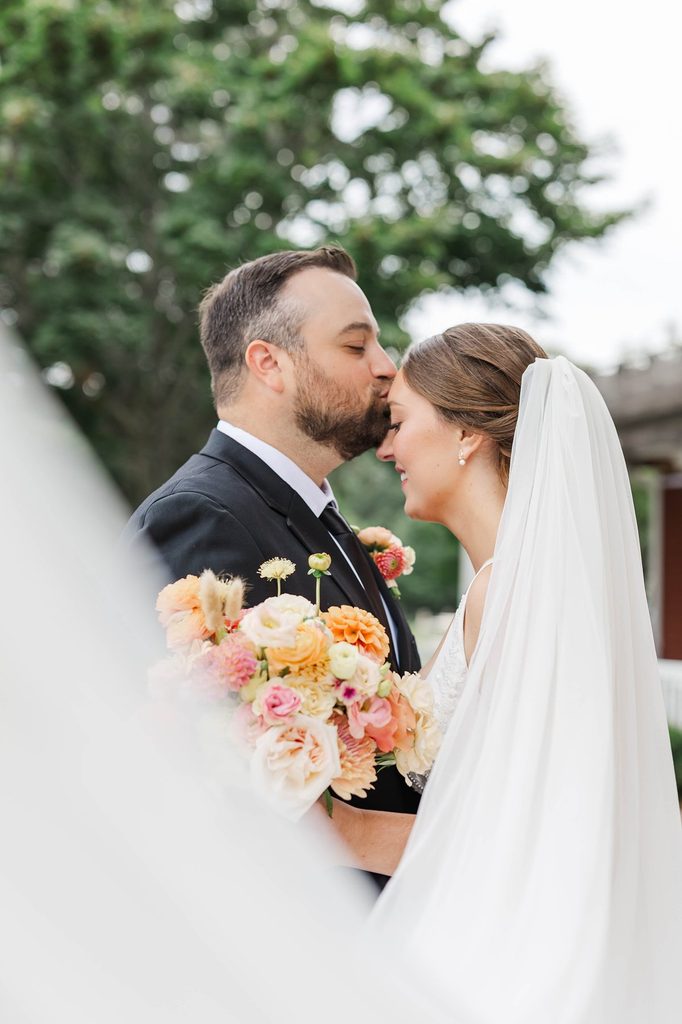 Cole kissing Allison on the forehead as she holds her colorful bridal bouquet during their wedding portraits at Black Star Farms.