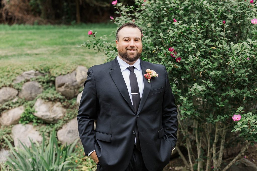 Portrait of Cole in his black wedding suit and boutonniere, standing outdoors among the greenery at Black Star Farms in Suttons Bay, Michigan.