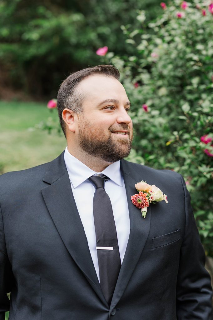 Natural wedding portrait of Cole smiling in his black suit with a colorful boutonniere against a garden backdrop at Black Star Farms.