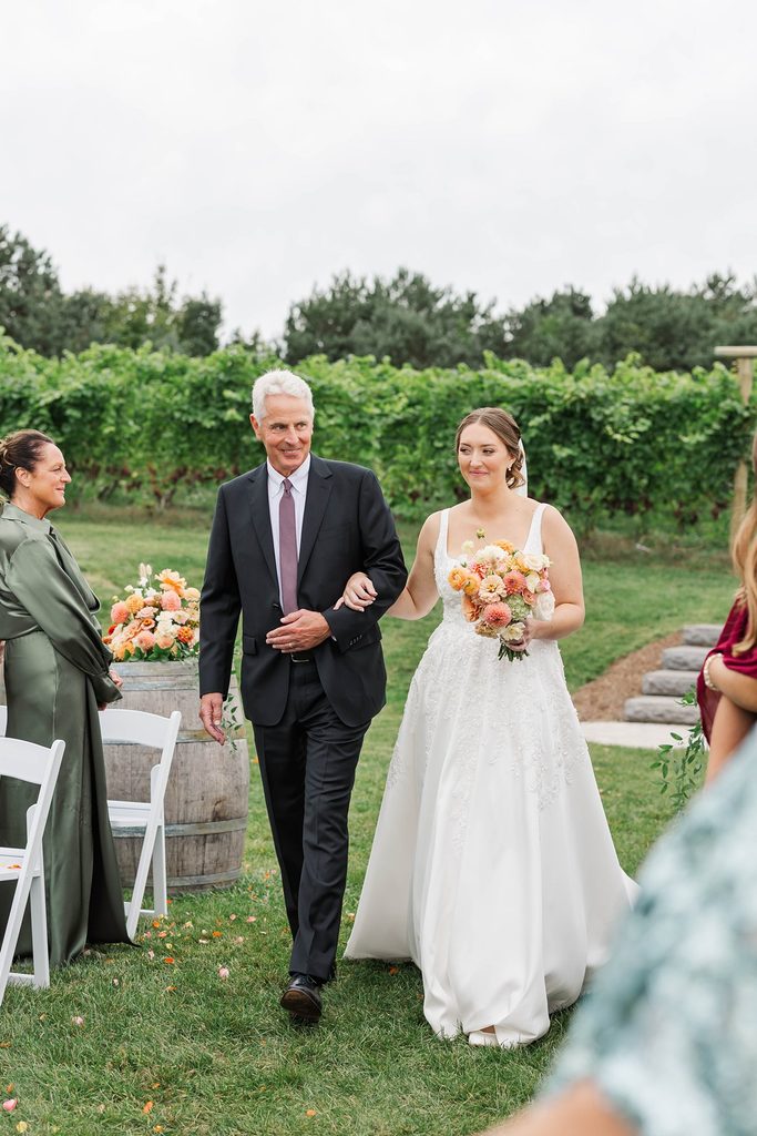 Allison walking down the aisle with her father, carrying a colorful bridal bouquet during their intimate vineyard wedding ceremony at Black Star Farms in Northern Michigan.