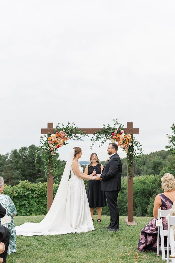 Allison and Cole exchanging vows beneath a floral-covered wooden arch during their intimate Black Star Farms vineyard wedding in Suttons Bay, Michigan.