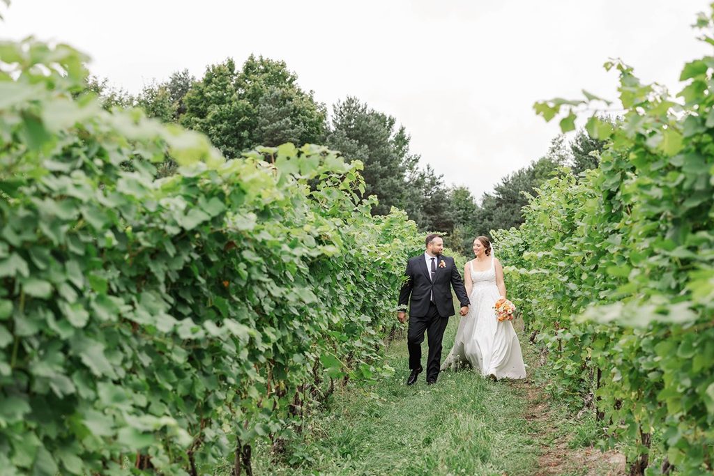 Allison and Cole walking hand in hand between lush grapevines during their Black Star Farms wedding in Suttons Bay, Michigan.