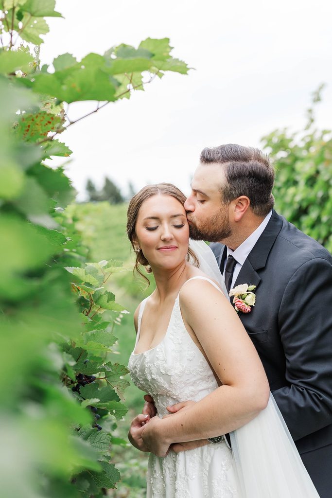 Cole gently kisses Allison's forehead between lush grapevines during their intimate Black Star Farms wedding in Northern Michigan.