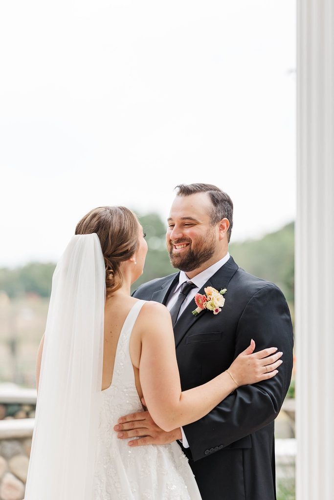 Cole smiling at Allison as they embrace during their romantic first look outside the Black Star Farms Inn in Suttons Bay, Michigan.