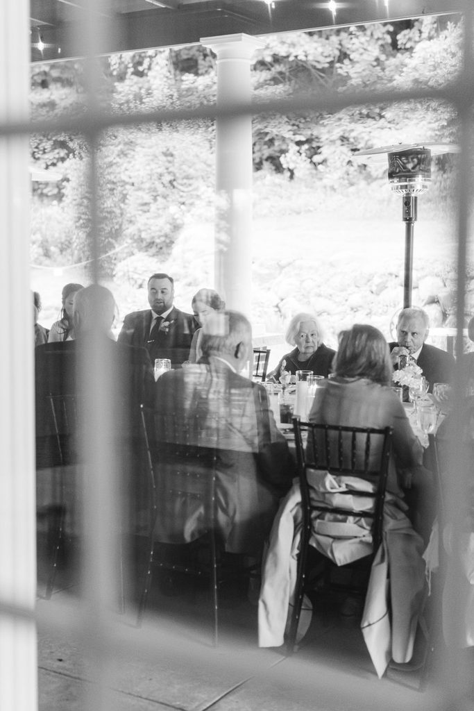 Candid black-and-white photograph of wedding guests gathered around the reception table beneath the covered patio at Black Star Farms winery.