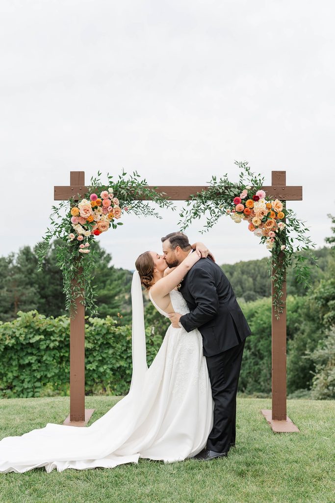 Allison and Cole share their first kiss as newlyweds beneath a colorful floral arch during their intimate Black Star Farms vineyard wedding in Suttons Bay, Michigan.